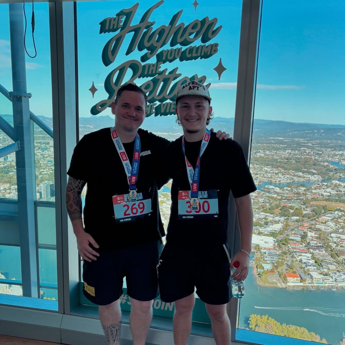 Tom and his apprentice Sam at the top of the Q1 tower wearing their climb numbers and finisher medals, with the Gold Coast behind them